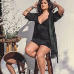 Woman in a black lingerie set posing for a professional portrait with a white background, sitting on a bench with flowers beside her.