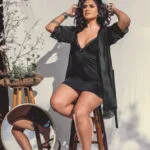Woman in a black lingerie set posing for a professional portrait with a white background, sitting on a bench with flowers beside her.