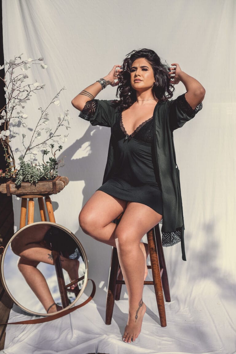 Woman in a black lingerie set posing for a professional portrait with a white background, sitting on a bench with flowers beside her.